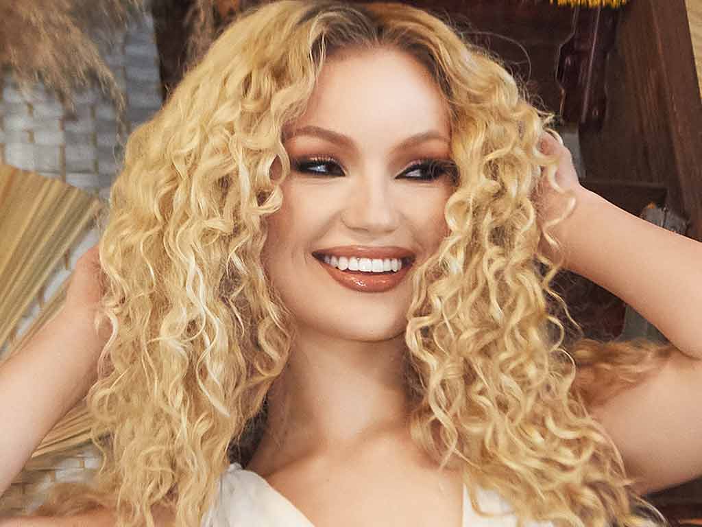 A woman with voluminous curly hair smiles brightly while holding her hands up to her head.
