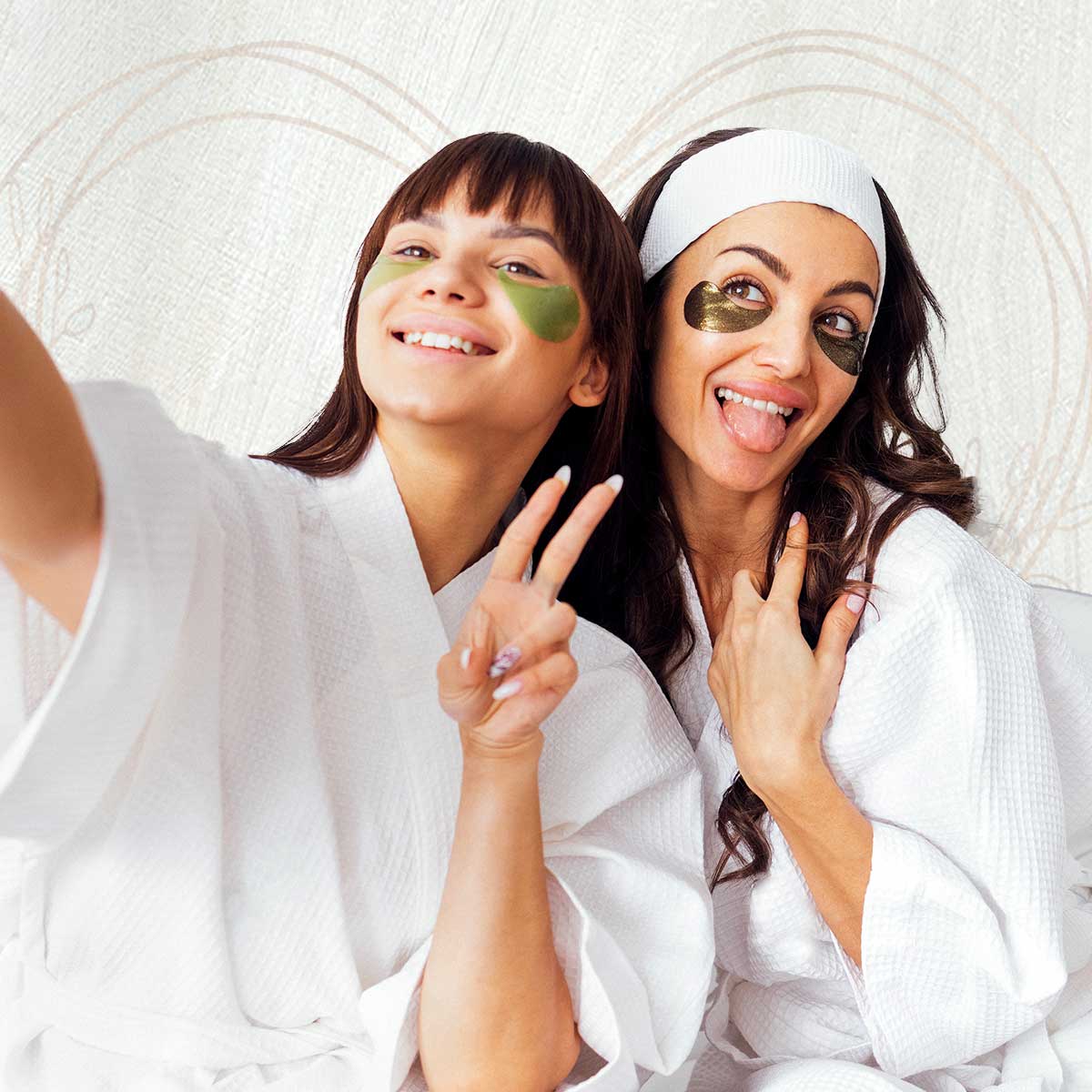 Two women in white robes and under-eye masks smile for a selfie during a spa day. One flashes a peace sign, the other sticks out her tongue playfully.