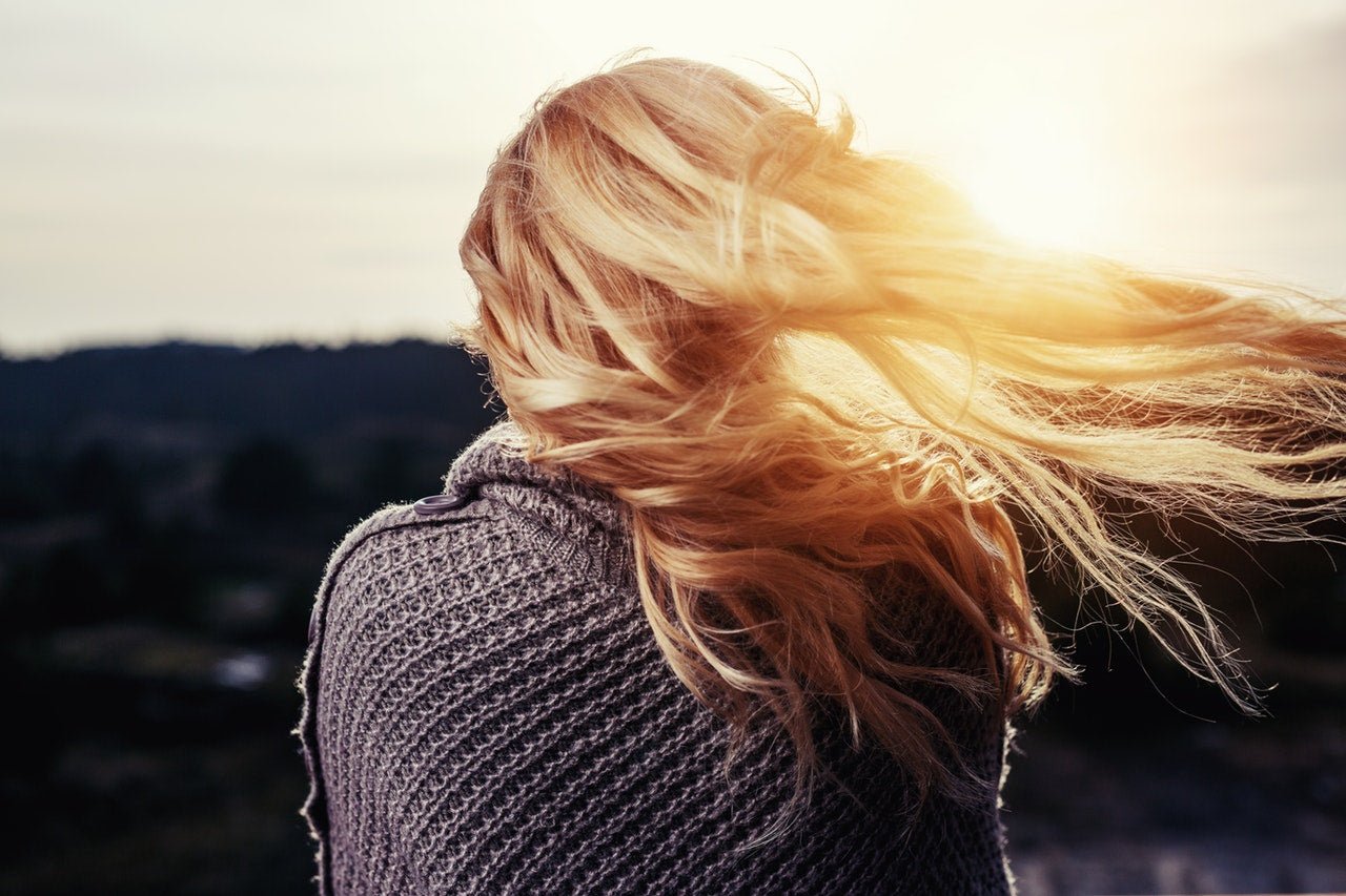 A person with long, blonde hair blowing in the wind is wrapped in a textured gray sweater, facing away from the camera toward a bright sunset, an image that captures the essence of winter hair care amid fading light.