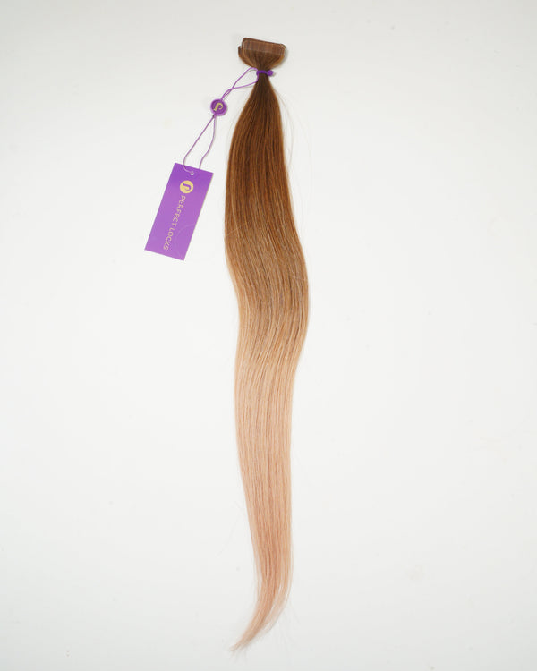 A single, long, ombre hair extension transitions from medium brown at the top to light blonde at the tip, attached to a purple tag with a string, set against a white background.