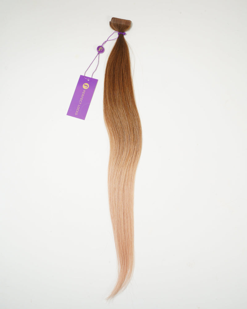 A single, long, ombre hair extension transitions from medium brown at the top to light blonde at the tip, attached to a purple tag with a string, set against a white background.