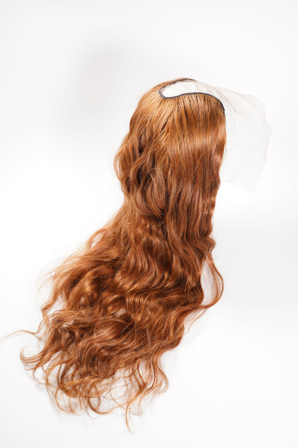 A long, wavy, reddish-brown lace front wig rests on a white surface, with loose curls cascading down.