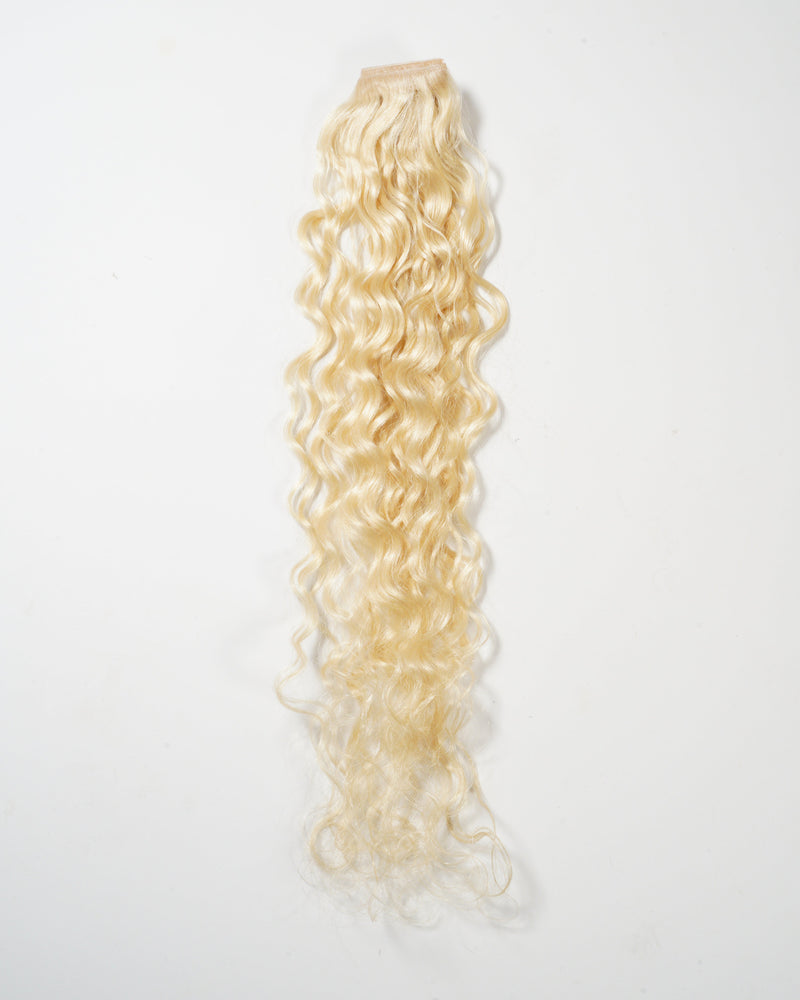 A long, blonde, curly hair extension with a sewn weft at the top is displayed on a white background, showcasing its voluminous texture.