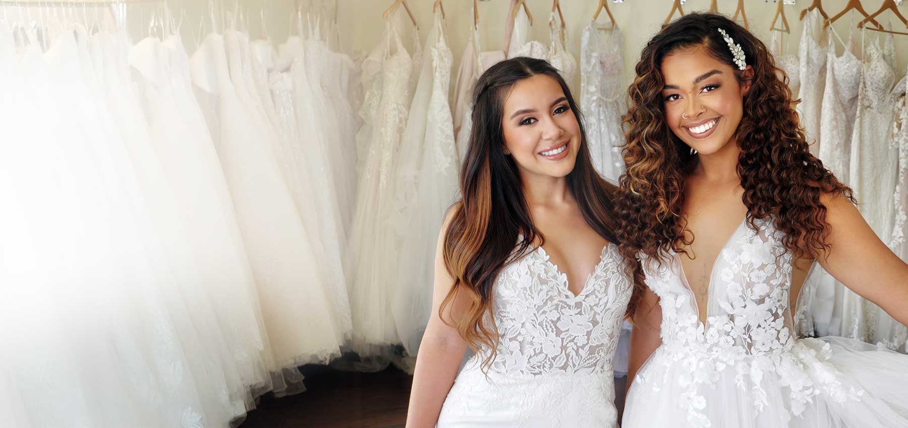 Two women in white bridal gowns stand smiling in a wedding dress shop, surrounded by elegant racks of white dresses on wooden hangers.