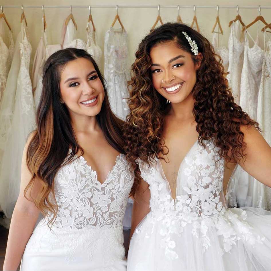 wo smiling brides in lace wedding gowns, one with long straight hair and one with long curly hair