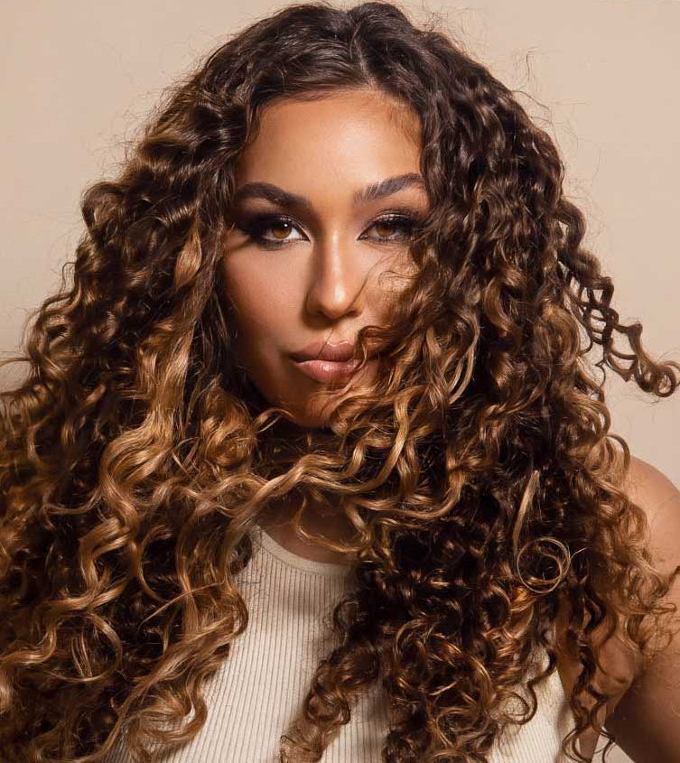 A close-up portrait of a woman with voluminous, tight curls in dark brown with warm caramel and golden highlights, wearing a cream ribbed top against a solid beige background.