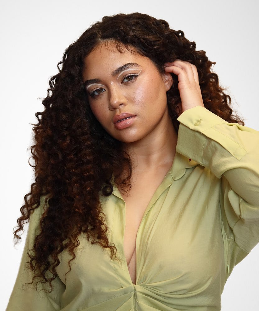 A woman poses confidently in a light yellow blouse, touching her hair styled with the Perfect Locks Tight Curly U-Part Wig in Chocolate Brown , set against a plain white background.#color_chocolate-brown-(3)background
