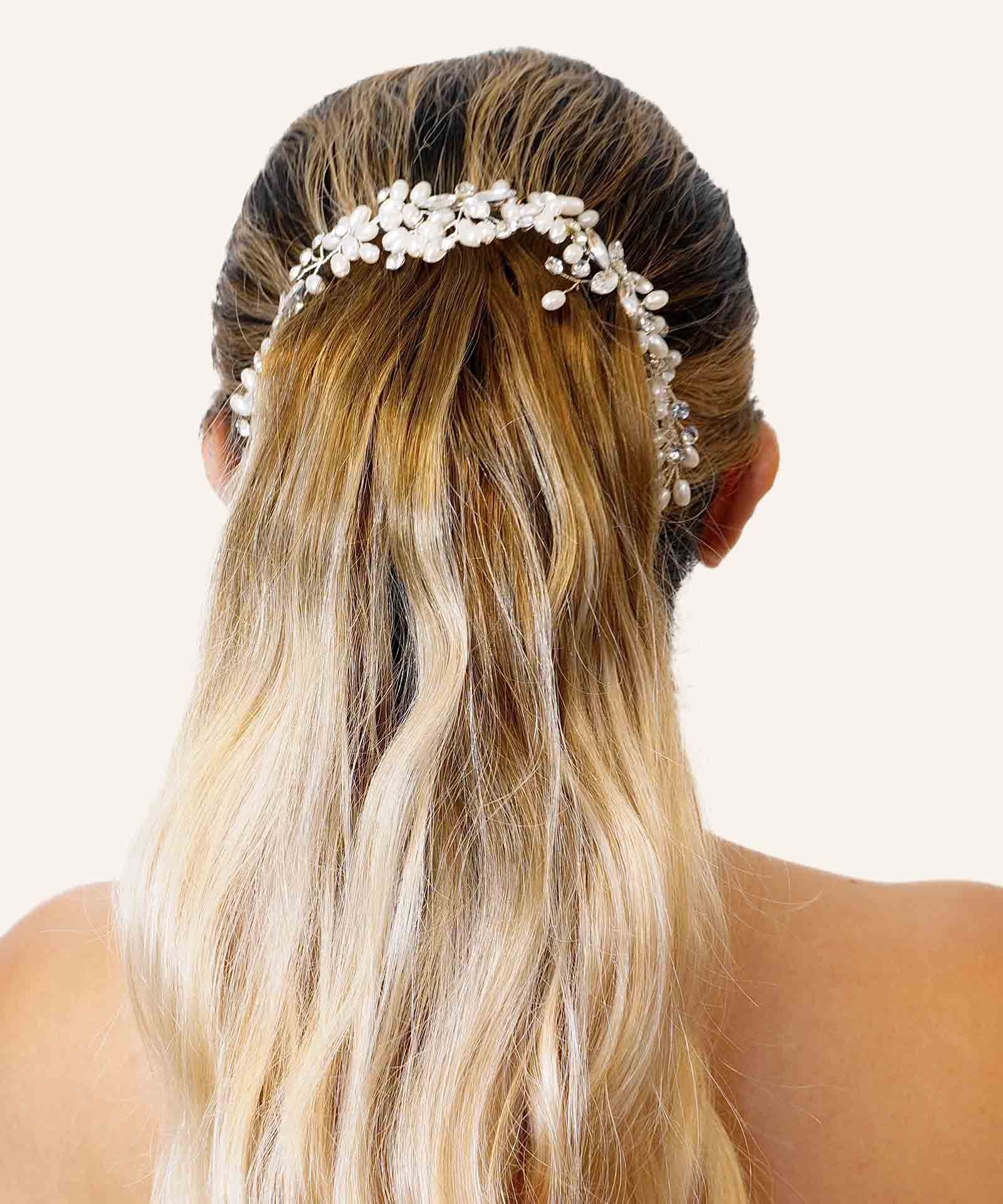 A woman with long, wavy blonde hair is seen from behind, wearing a delicate white flower and pearl hair accessory. Plain background.