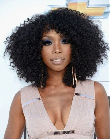 A woman with voluminous curly hair styled with kinky curly extensions, dramatic dark eye makeup, and nude lipstick wears a sleeveless beige dress with metallic accents, posing in front of a light background with a geometric design.