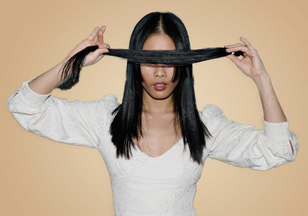A woman with long black hair, channeling Hair Myths, wears a white blouse and holds a section of her hair horizontally across her eyes, covering them, against a plain beige background.