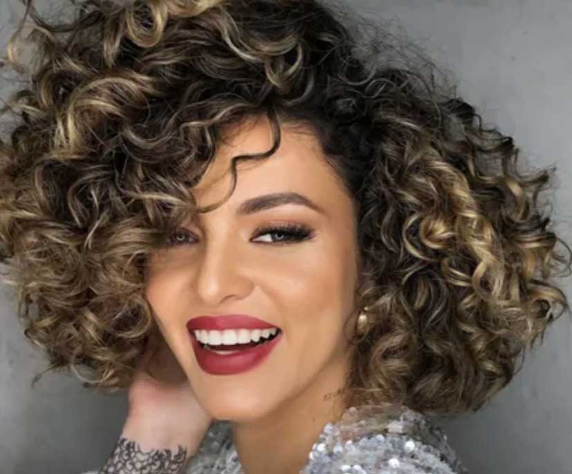Woman with short, voluminous curly hair and blonde highlights smiling confidently, wearing red lipstick and a silver sequin top against a gray background.