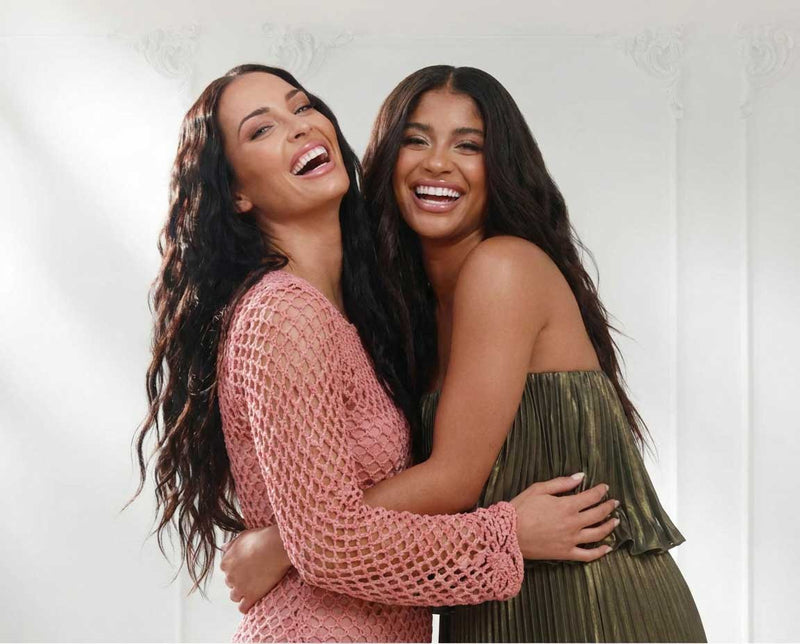 Two women with long, wavy hair are smiling and hugging each other in a bright room. One wears a pink, netted dress, and the other wears a strapless, green pleated dress. They look happy and joyful.