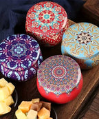 Four round tins with vibrant mandala designs rest on a wooden surface, beside small bowls of assorted cubed cheese and dried fruit.