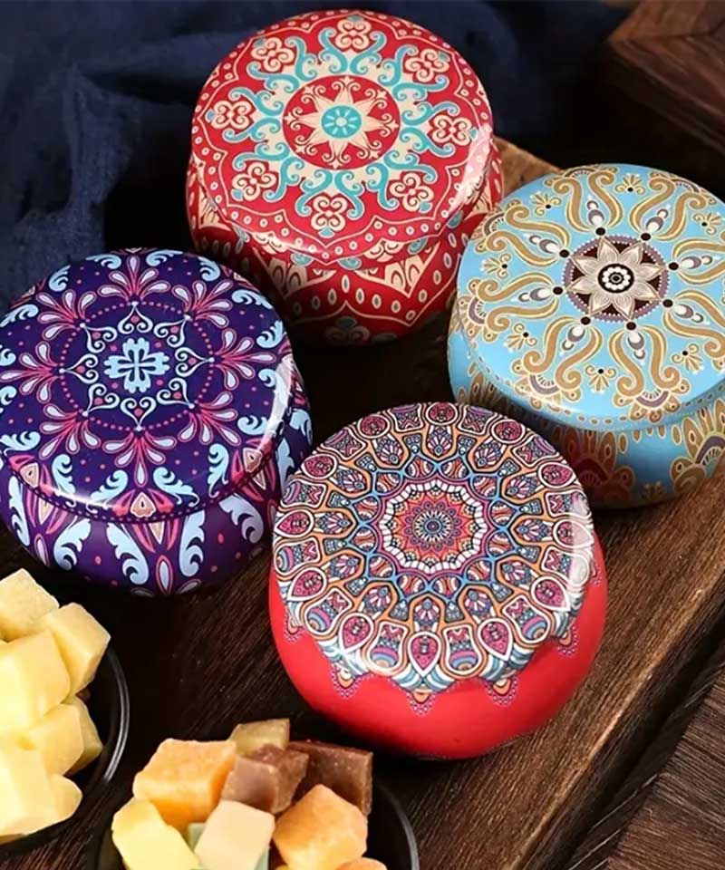 Four round tins with vibrant mandala designs rest on a wooden surface, beside small bowls of assorted cubed cheese and dried fruit.