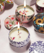 Several decorative tin candles with floral patterns are lit on a light pink surface, matching lids nearby, each uniquely designed.
