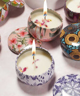 Several decorative tin candles with floral patterns are lit on a light pink surface, matching lids nearby, each uniquely designed.