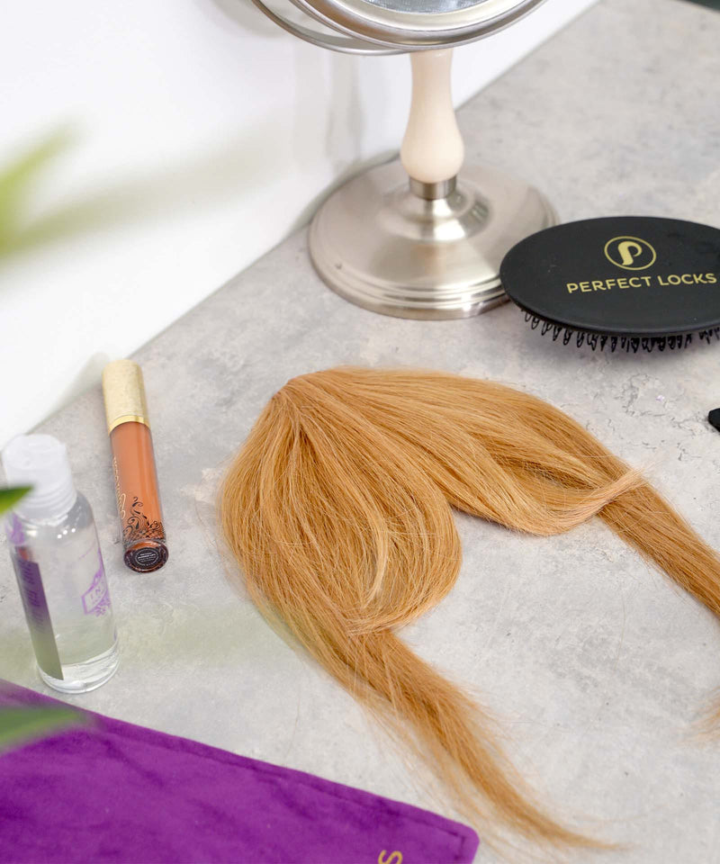 A blond hair extension rests on a gray countertop beside a hairbrush, makeup mirror, lip gloss, spray bottle, and purple cloth.