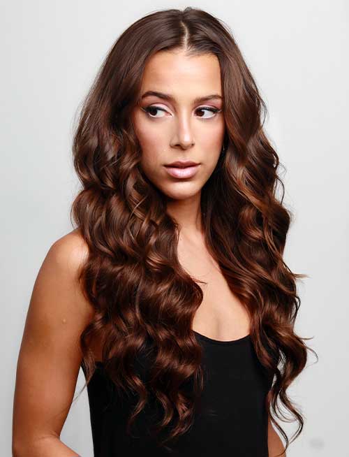 A woman with long, voluminous, chocolate brown waves wearing a black sleeveless top gazes to her left against a simple, light background.