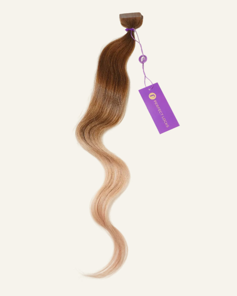 A long, wavy hair extension in brown to blonde ombre, tied at the top with a purple string and matching tag. This stylish hairpiece is showcased against a plain, light background.