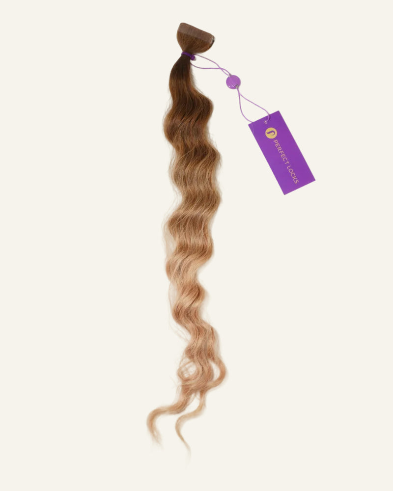 Wavy hair extensions with a brown-to-blonde ombre color, held together at the top by a band and tagged with a purple label, are displayed on a light background.