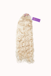 A bundle of long, wavy, platinum blonde hair extensions with a purple tag at the top on a plain white background.