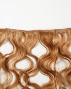 A section of wavy, reddish-brown hair extensions is displayed against a white background, highlighting the texture and soft waves of the strands.