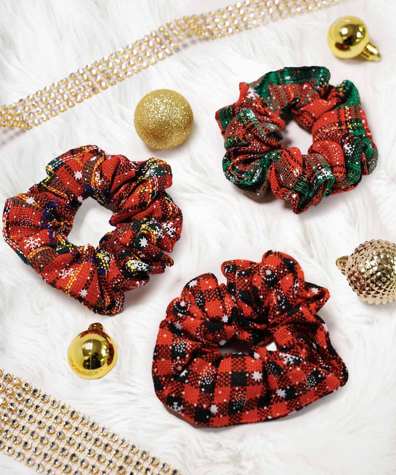 Three festive scrunchies with red and green patterns on a white fluffy surface, surrounded by gold ornaments and sparkly ribbon.