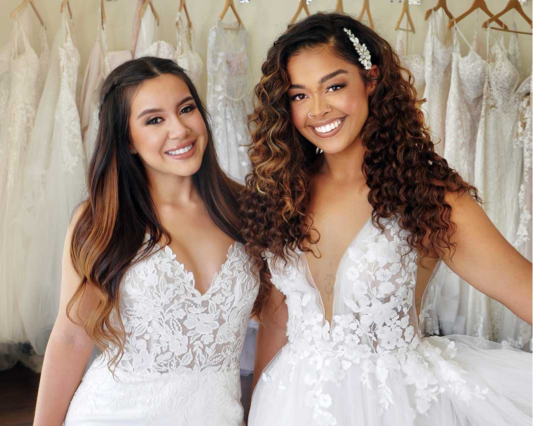 wo smiling brides in lace wedding gowns, one with long straight hair and one with long curly hair