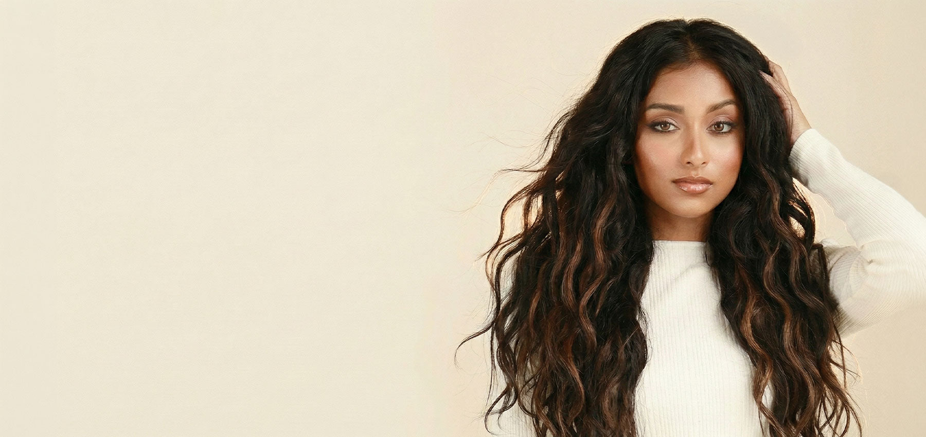 Woman with long voluminous dark wavy hair extensions touching her hair, wearing a white top against a warm cream background, showcasing Perfect Locks Remy human hair extensions
