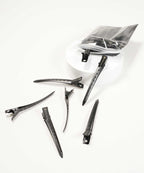 Six black hair clips scattered on a white surface near a round container and a partially open clear plastic bag with more clips.