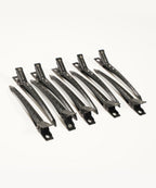 Six black hair sectioning clips are arranged in two neat rows on a white background, each with holes and a secure spring mechanism.
