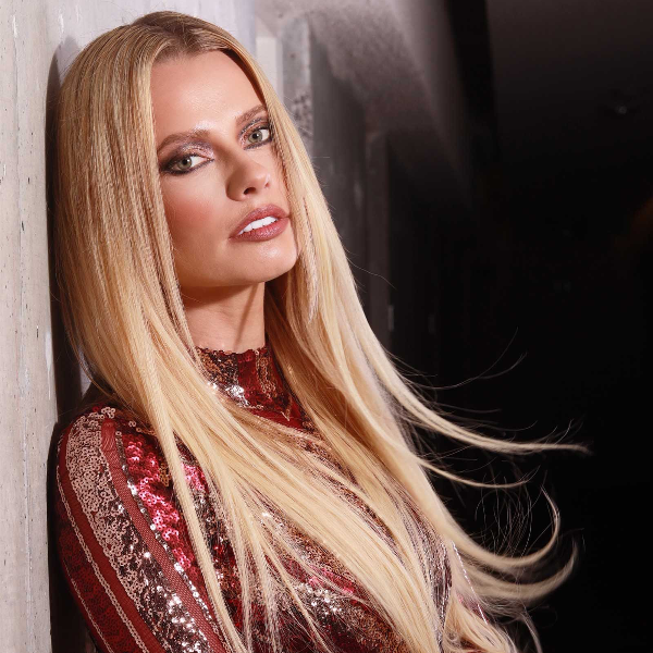 Woman with long blonde hair wearing a red and gold sequined outfit against a dark background