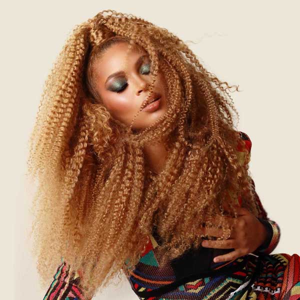 Woman with long, voluminous honey-blonde textured curly hair extensions posing confidently with soft glam makeup.