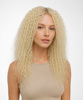 A woman with long, voluminous, tight curls in a Platinum Blonde wig wears a sleeveless beige top against a plain white background.#color_platinum-blonde-(613)