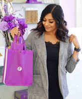 A woman with wavy dark hair in a plaid blazer and black top smiles, holding a purple gift bag. Purple flowers decorate the background.