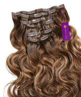 Several wefts of wavy Seamless Clip-In Hair Extensions in rooted toffee blonde highlights are neatly arranged with attached clips and a purple Perfect Locks tag on the right.#color_rooted-toffee-blonde-highlights-(4/27/4)