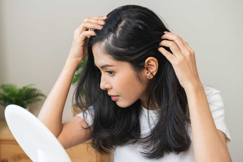 A woman with long dark hair and a white shirt examines her scalp in a mirror, parting her hair. A blurred plant is in the background.