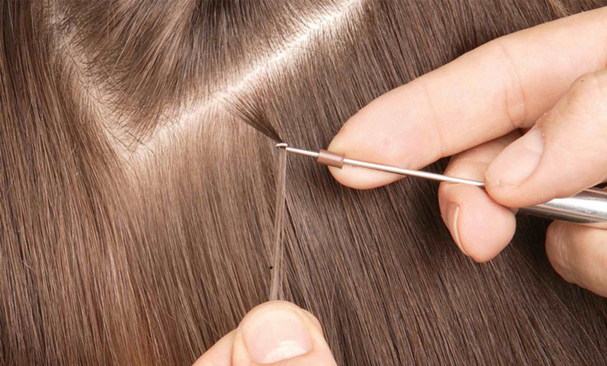 Hands skillfully use a hook tool to install seamless hair extensions on straight, light brown hair near the scalp.