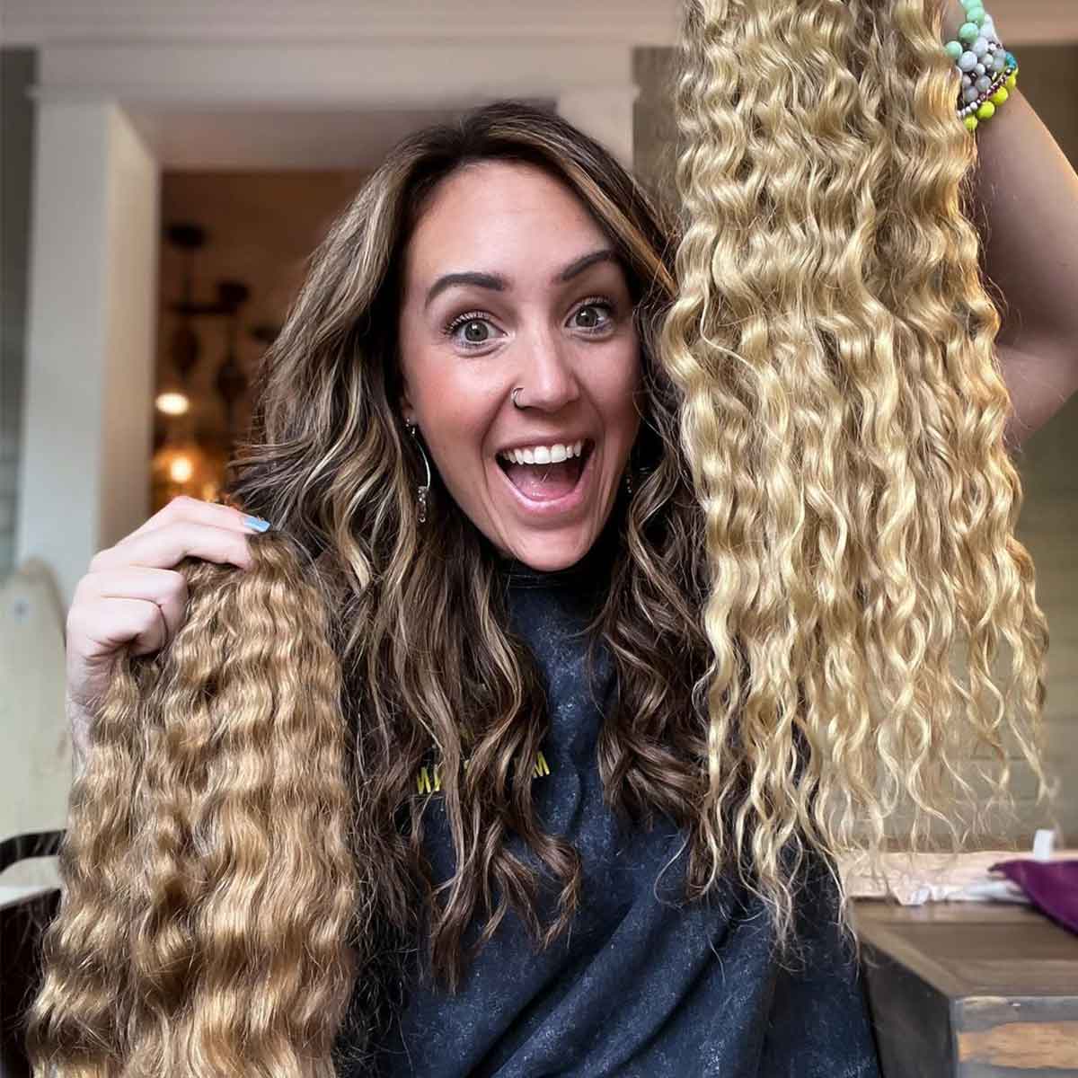 A smiling woman with wavy brown hair holds up two long, wavy blonde hair extensions in a warmly lit room, showcasing their vibrant color and texture.