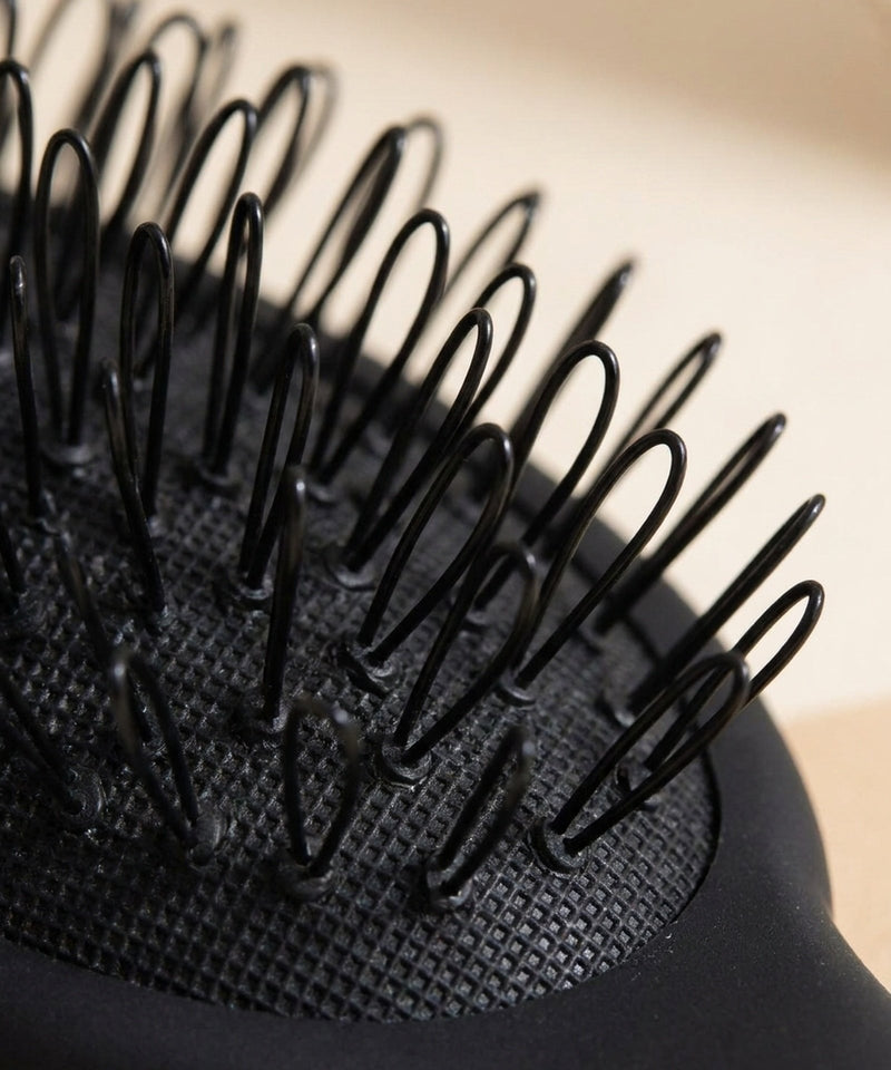 Matte black hair extension brush showing curved wire loop bristles on a textured cushion against a neutral background.