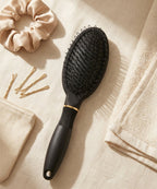 Matte-black loop hair brush with gold trim on beige linen alongside a silk scrunchie and gold bobby pins.
