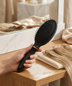 Hand holding a matte black loop hairbrush with gold trim beside blonde weft extensions on a wooden vanity table.