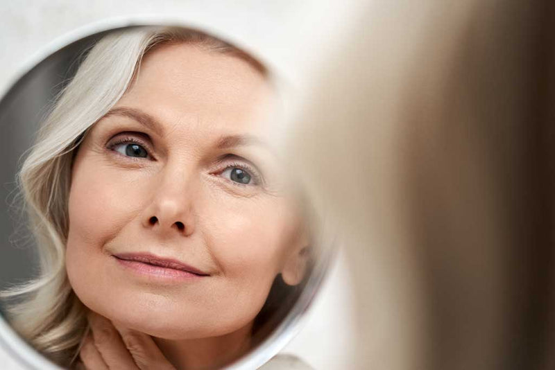 A mature woman with light hair admires her reflection in a round mirror, gently touching her chin and smiling softly.