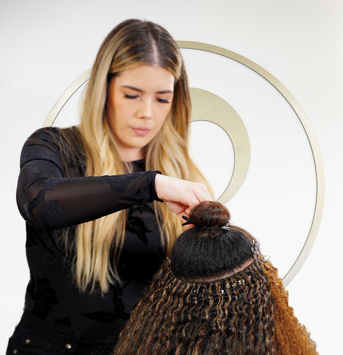 A hairstylist with long blonde hair seamlessly attaches extensions to a client's curly hair, set against a decorative circular pattern.