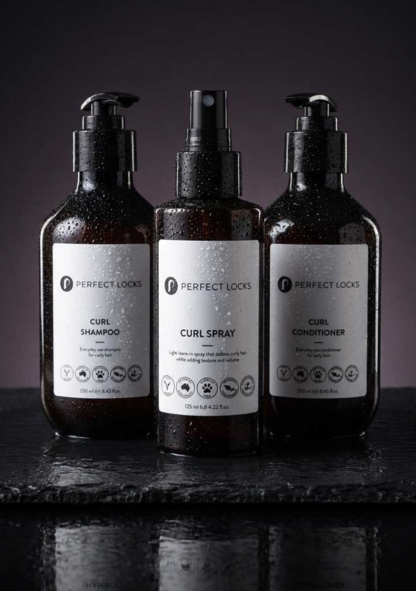 Three Perfect Locks curl care bottles - shampoo, spray, and conditioner - with water droplets on dark slate