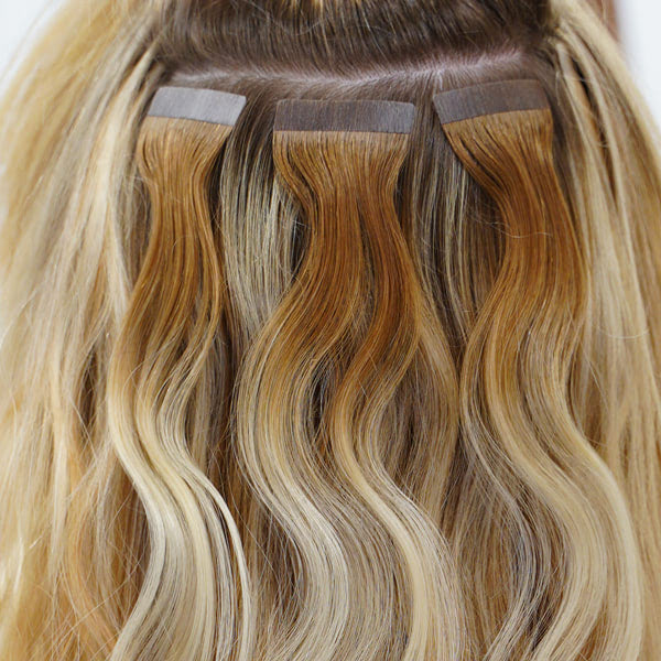 Close-up of blonde hair with three wavy hair extension strips near the roots, showing the seamless tape-in method for natural volume.