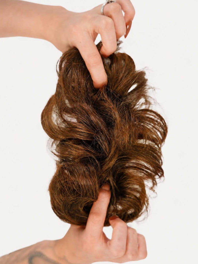 Rich Brunette (2B) human hair scrunchie extension stretched between two hands, featuring dimensional medium brown tones with warm caramel highlights and soft wavy texture on a clean light background.