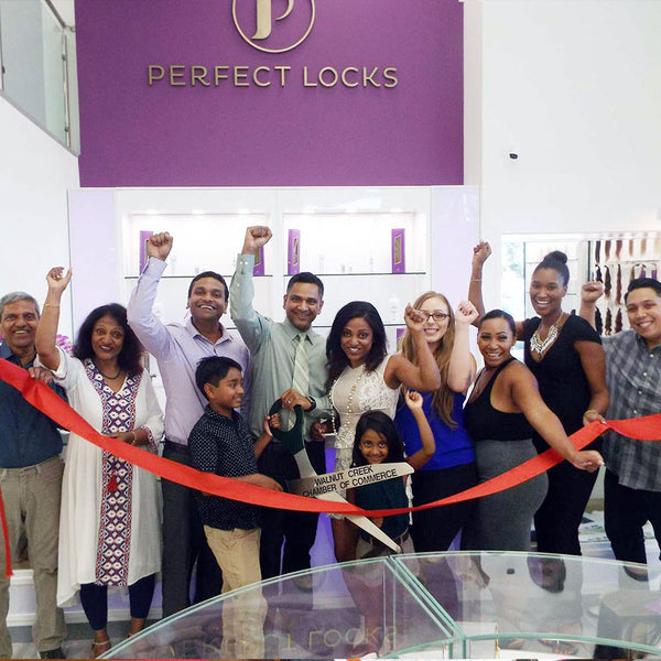 A diverse group smiles and raises arms at a ribbon-cutting inside Perfect Locks, as children hold a red ribbon for the grand opening.