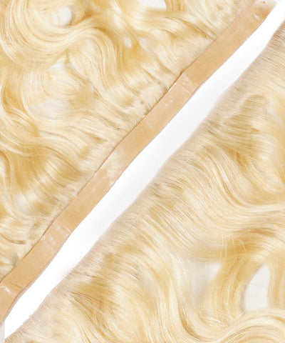 Close-up view of wavy Seamless Tape In Weft extensions, light blonde Remy hair with beige keratin adhesive on white.