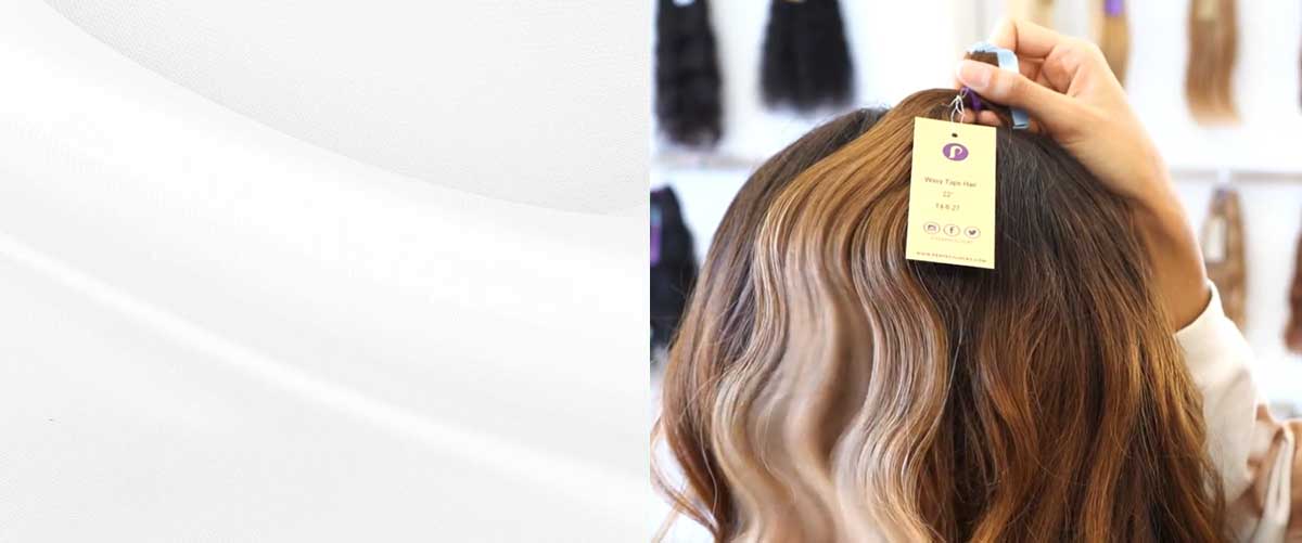 A person displays a bundle of wavy brown hair extensions with a tag attached, set against a background of hanging hairpieces.